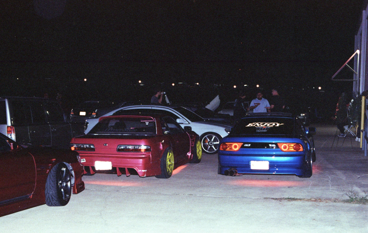 A nighttime scene showing several cars parked in a lot, with Pulse's pink S13 Silvia in the center-left and a blue car in the center-right. The blue car has 'ENJOY' written on the back window.