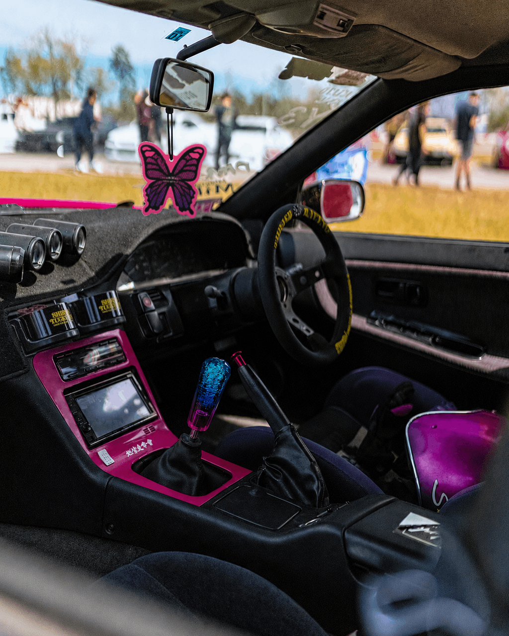 Close-up of a customized car interior with a black dashboard and center console, bright pink trim around the glovebox and center console, a manual gear shifter with a blue-tinted knob, a black steering wheel, and a butterfly-shaped air freshener hanging in the windshield. A person is seated in the driver's seat, and the car is photographed outdoors with blurred people and vehicles visible through the windshield.