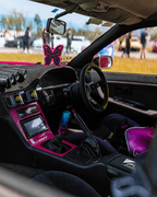 Close-up of a customized car interior with a black dashboard and center console, bright pink trim around the glovebox and center console, a manual gear shifter with a blue-tinted knob, a black steering wheel, and a butterfly-shaped air freshener hanging in the windshield. A person is seated in the driver's seat, and the car is photographed outdoors with blurred people and vehicles visible through the windshield.