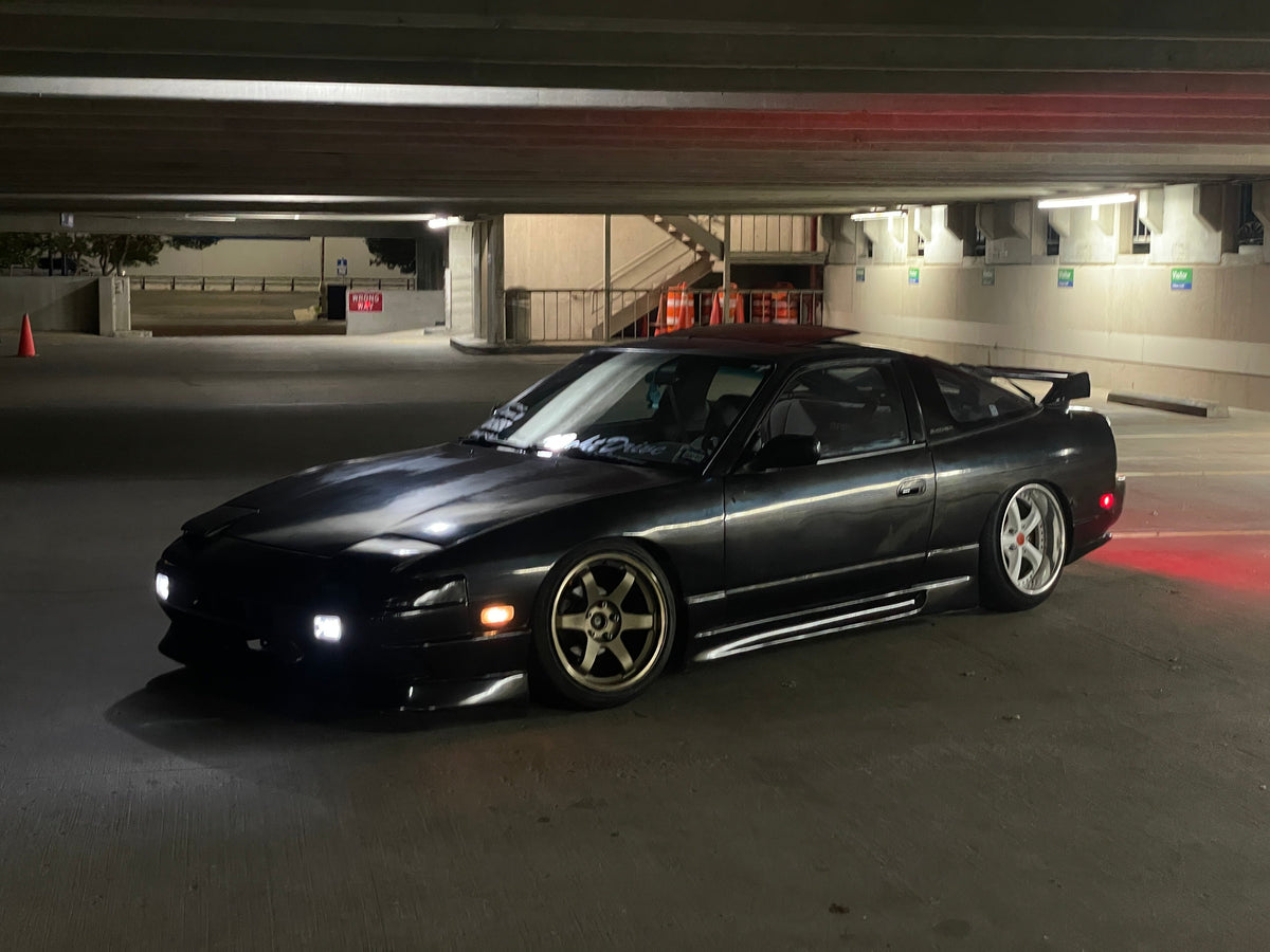 A dark metallic gray Nissan 180SX sports car with custom gold multi-spoke wheels, parked in a dimly lit parking structure at night.