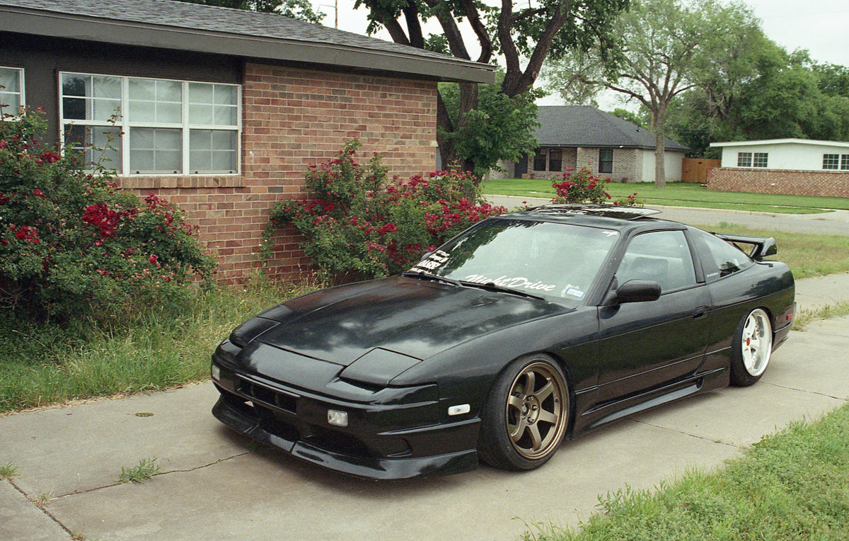 A black modified Nissan 240SX with a front splitter, rear spoiler, and bronze multi-spoke wheels, parked on a concrete driveway in front of a brick house.