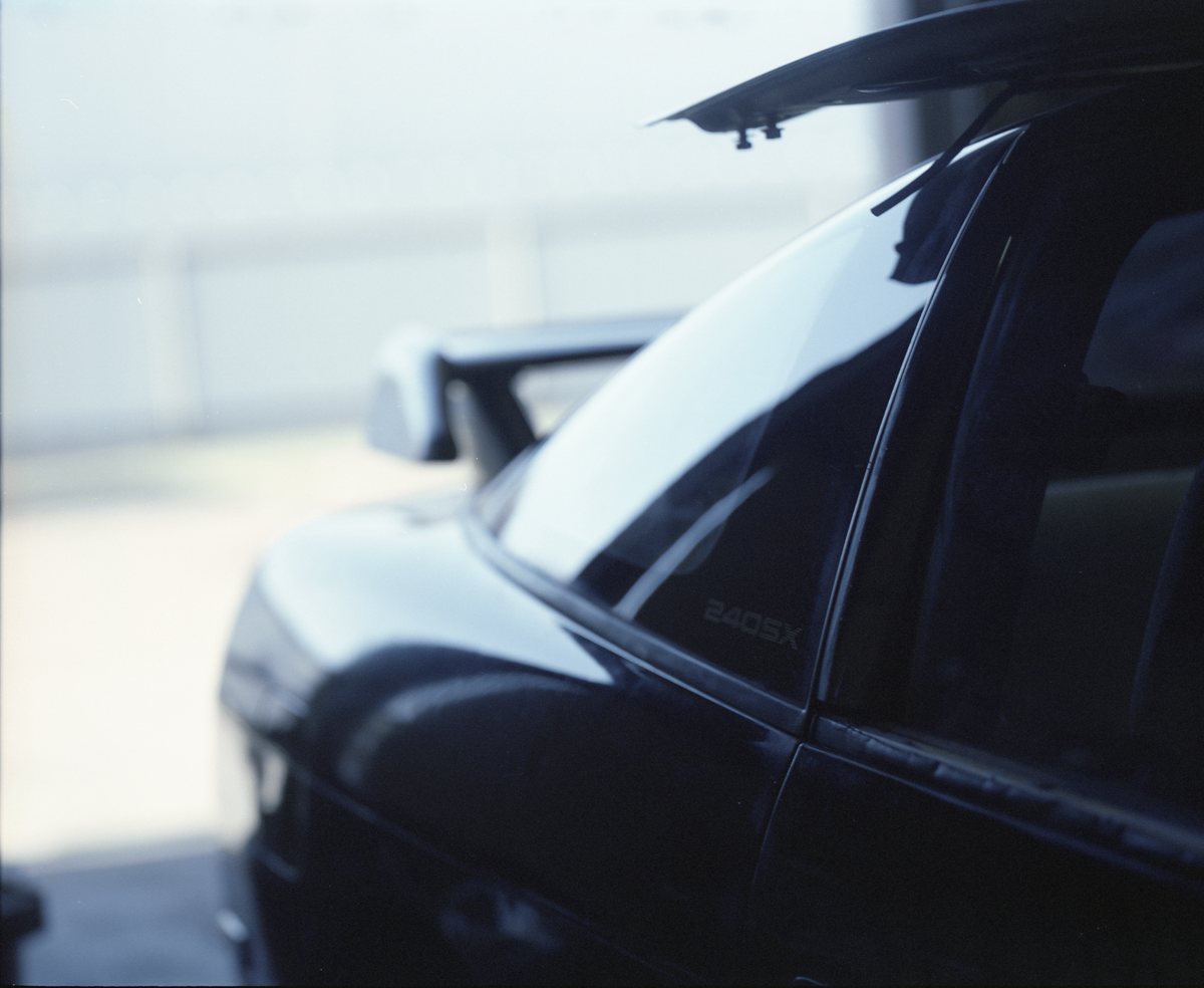 Close-up side view of a glossy black car with a small '240SX' badge on the window, photographed outdoors with a blurred light gray background.