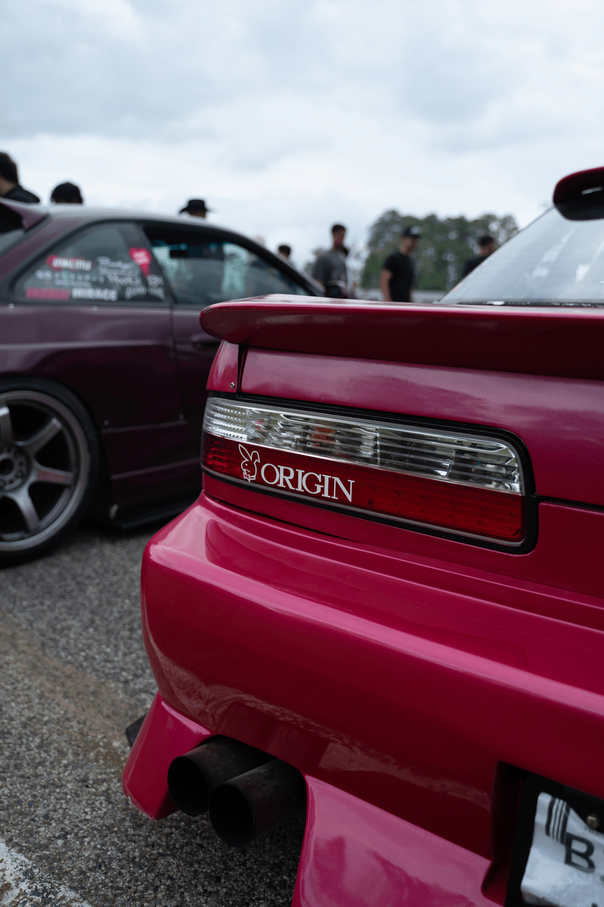 Close-up rear view of a bright pink Nissan Silvia S15 with a custom red rear bumper and a visible 'ORIGIN' badge, parked outdoors at an event with another car in the background.