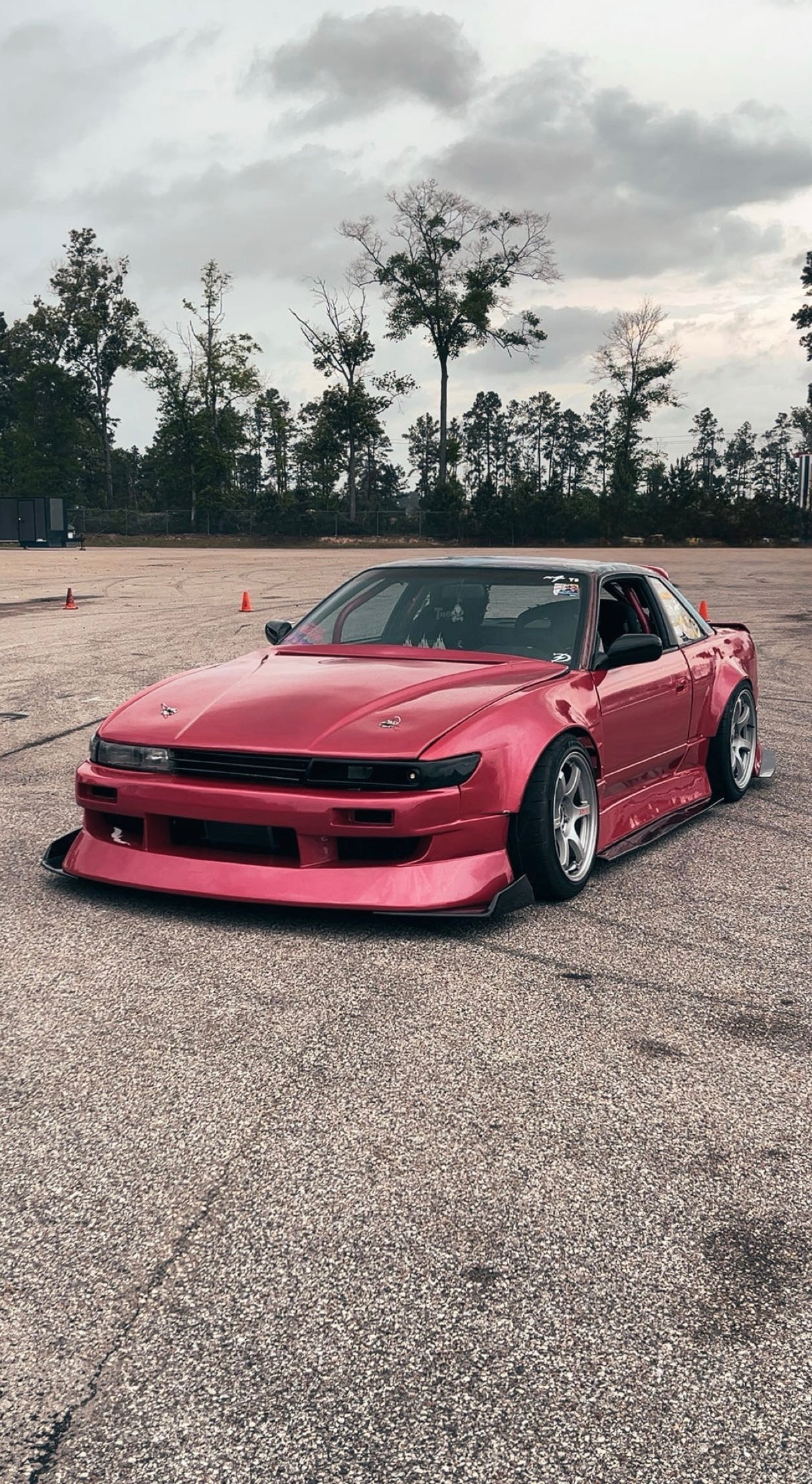 A pink widebody Nissan S13 Silvia with a lowered suspension and Origin Drift Line kit, parked on an asphalt lot with trees in the background under a cloudy sky.