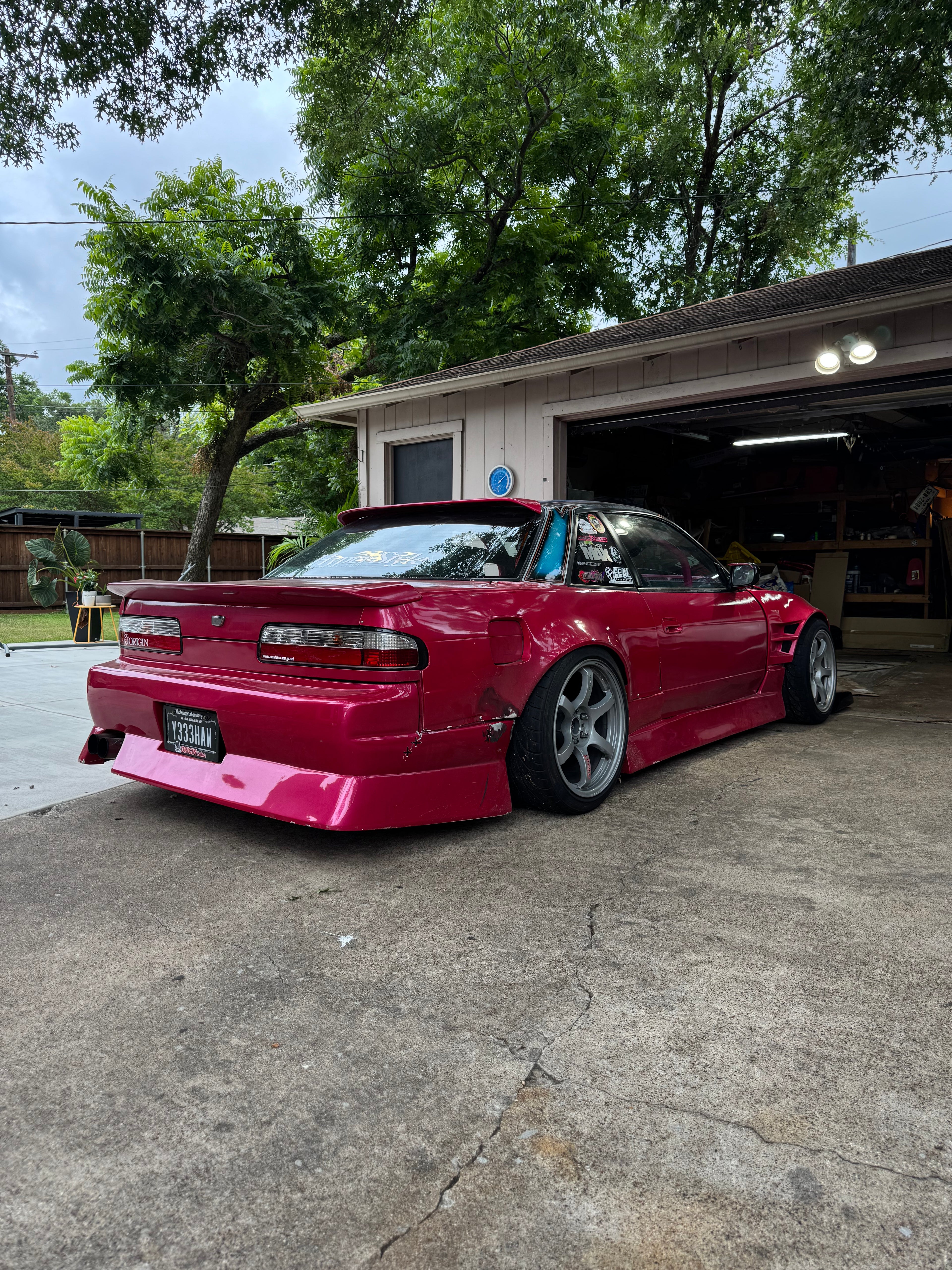 A bright pink customized Nissan S13 Silvia coupe with widebody fenders and a lowered suspension, parked in a residential driveway in front of a garage.