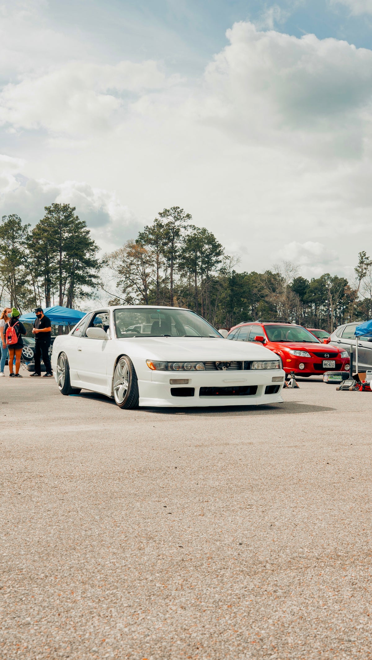 A white Nissan S13 Silvia parked on an outdoor paved lot at a car show, with other cars, trees, and people visible in the background under a cloudy sky.