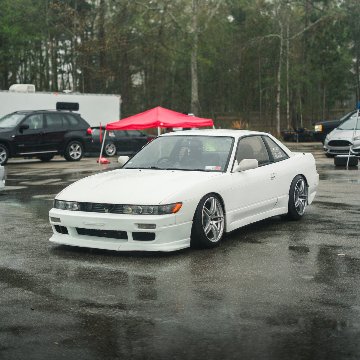 A white Nissan S13 Silvia coupe parked on a wet asphalt lot with other vehicles, a red canopy, and trees in the background.